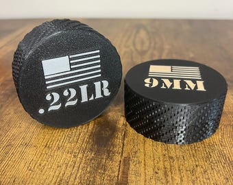 May include: Two black, cylindrical objects with textured sides. One has the text ".22LB" and an American flag graphic. The other has "9MM" and a similar flag design. Both are on a wooden surface.