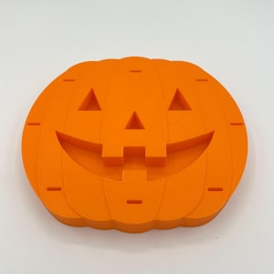 May include: A bright orange, flat, pumpkin-shaped object with a carved jack-o'-lantern face. The design features two triangular eyes, a triangular nose, and a wide, curved mouth with two square teeth. The surface has vertical ridges.