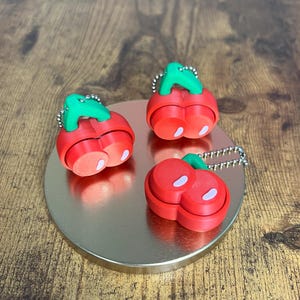 May include: Three red cherry-shaped keychains with green stems and white accents. Each keychain is attached to a silver chain. The keychains are arranged on a gold-colored, circular surface.