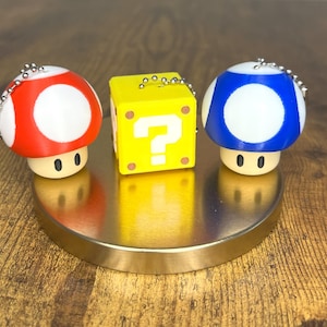 May include: Three keychains featuring characters from a video game. A red mushroom with white spots, a yellow question mark block, and a blue mushroom with white spots. Each keychain has a silver chain and is on a gold-colored stand.