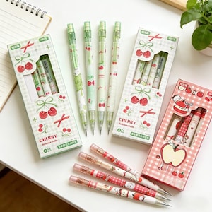 May include: A selection of decorative mechanical pencils and pencil boxes. The pencils feature cherry and apple designs in red, green, and white. The boxes are decorated with cherry and apple graphics. The text "CHERRY delicious fruits" is visible on the boxes.