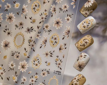 May include: Clear sheet of nail art stickers featuring various floral designs in shades of brown and white, with gold accents. The sheet includes flowers, leaves, and ornate frames. Several decorated nails are displayed, showcasing the sticker designs.