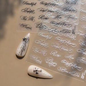 Könnte beinhalten: Nagelkunst-Aufkleber mit elegantem Schreibschrift-Text, darunter Wörter wie "Beautiful", "Rose" und "Twilight Whispers". Gezeigt werden zwei künstliche Nägel, einer mit einem Stern und der andere mit einem Kreuz-Charm.