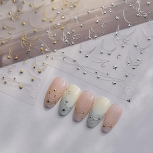 May include: Nail art stickers featuring gold and silver celestial designs of stars and crescent moons. The stickers are applied to manicured nails in various pastel colors, including pink, white, and blue. The nail art stickers are on a clear sheet.