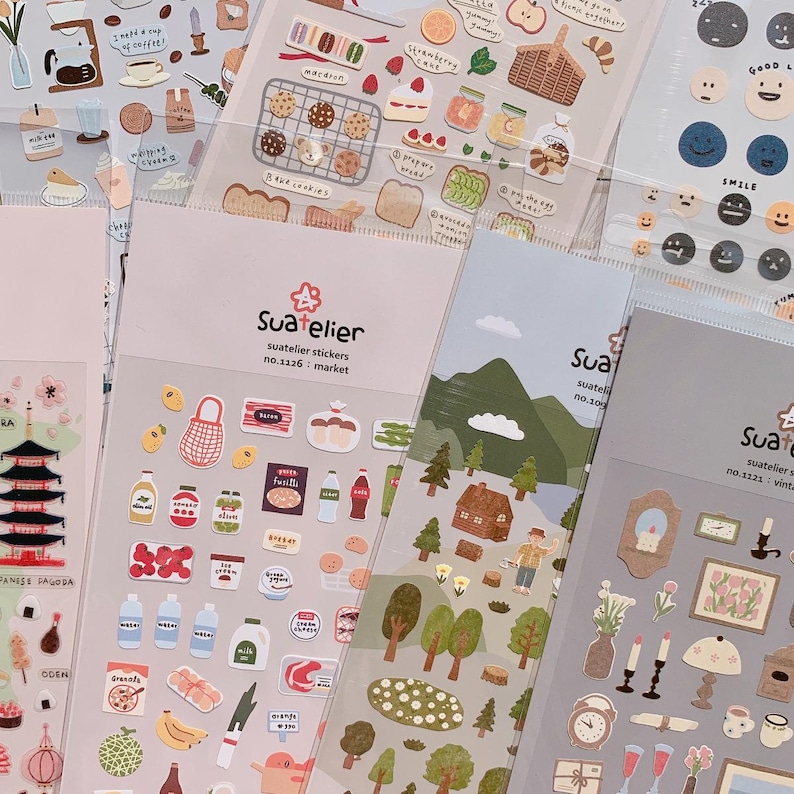 May include: A collection of Suatelier sticker sheets featuring various designs. The stickers include food items like macarons, bread, and fruit, along with nature scenes and vintage-themed illustrations. The text "Suatelier" and "market" are visible.