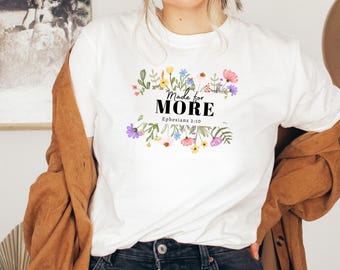 May include: White t-shirt with the text "Made for MORE" in black, surrounded by a floral design in various colors. The text "Ephesians 2:10" is below the main text. The shirt is a casual wear item.