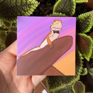 May include: A cartoon illustration of a bearded dragon wearing a black bow tie and sitting on a brown branch against a purple and orange sunset background.