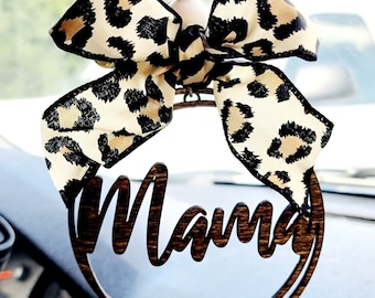 Peut inclure: Un porte-bonheur de voiture en bois avec le mot "Mama" écrit en cursive. Le porte-bonheur est décoré d'un nœud en imprimé léopard brun et noir et de perles en bois.
