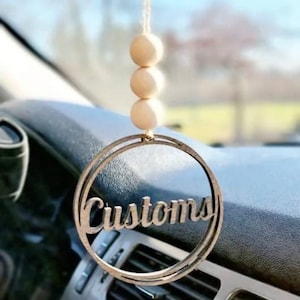 May include: A wooden car charm with the word "Customs" in a circle. The charm is hanging from a rearview mirror with three wooden beads.