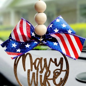 May include: A wooden car charm with the words "Navy Wife" in a circle. The charm is decorated with a red, white, and blue bow and three wooden beads.