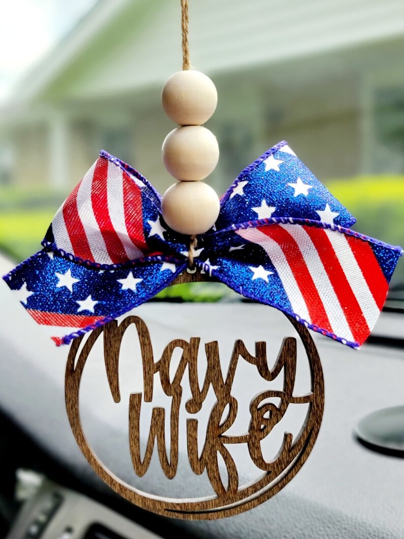 May include: A wooden car charm with the words "Navy Wife" in a circle. The charm is decorated with a red, white, and blue bow and three wooden beads.