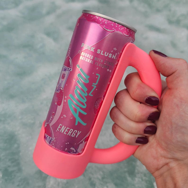 May include: A pink energy drink can with the text "Pink Slush" and "Alani Nu Energy" is held in a bright pink handle. The can features a strawberry and ice cream graphic. The handle is designed to fit around the can for easy gripping.