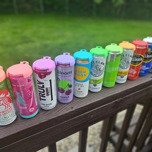 May include: A row of beverage cans with colorful lids, including Starbucks, Truly, and White Claw. The cans are arranged on a wooden railing against a green background. The lids are in various colors, such as pink, purple, and green.