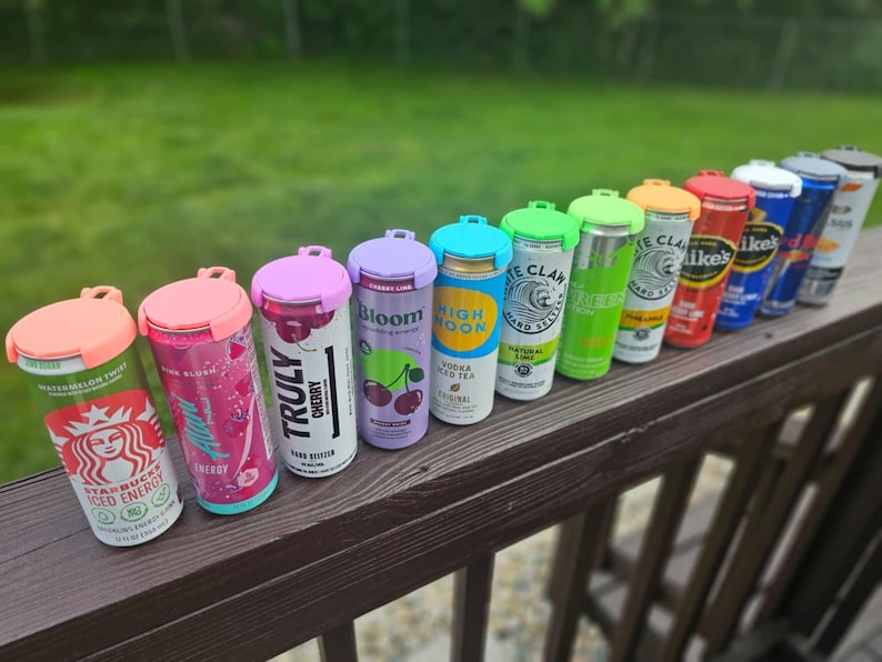 May include: A row of beverage cans with colorful lids, including Starbucks, Truly, and White Claw. The cans are arranged on a wooden railing against a green background. The lids are in various colors, such as pink, purple, and green.