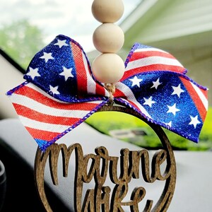 May include: A wooden car charm with the words "Marine Wife" in a circle. The charm is decorated with a red, white, and blue bow and wooden beads.