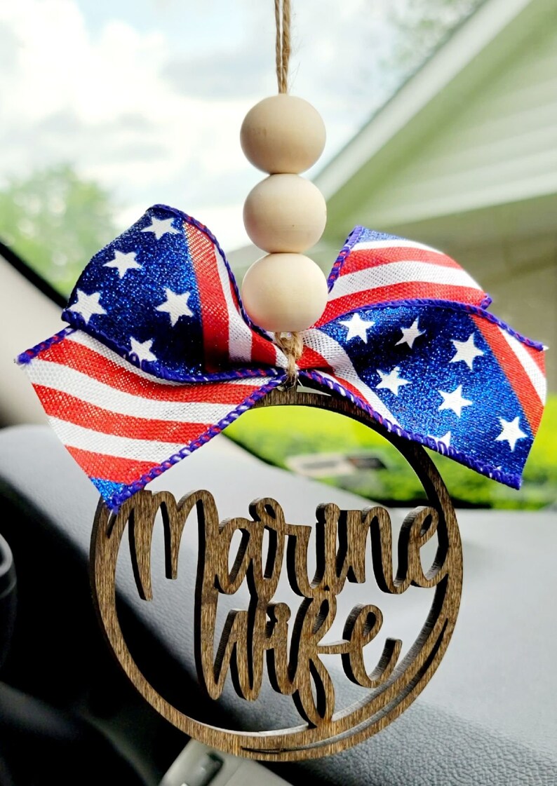May include: A wooden car charm with the words "Marine Wife" in a circle. The charm is decorated with a red, white, and blue bow and wooden beads.