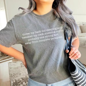 May include: A gray t-shirt with a white graphic print that reads "threw my back out sleeping and tweaked my neck sneezing so I'm probably one strong fart away from complete paralysis."