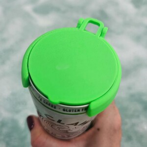 May include: A bright green can lid with a handle is attached to a can. The can is white with black text and a swirl design. The lid is made of plastic and is designed to fit snugly on the can.