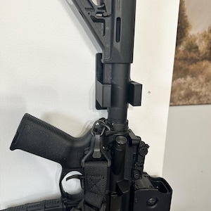 May include: A black rifle with a black sling and a black scope mounted on top. The rifle is mounted on a wall with a black wall mount. The rifle has a black magazine attached to the bottom. The magazine has a sticker on it that says "POSSIBLY BLANKET".