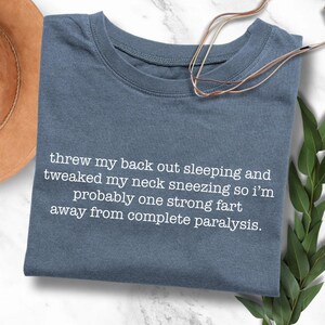 May include: A blue t-shirt with white text that reads "threw my back out sleeping and tweaked my neck sneezing so i'm probably one strong fart away from complete paralysis."