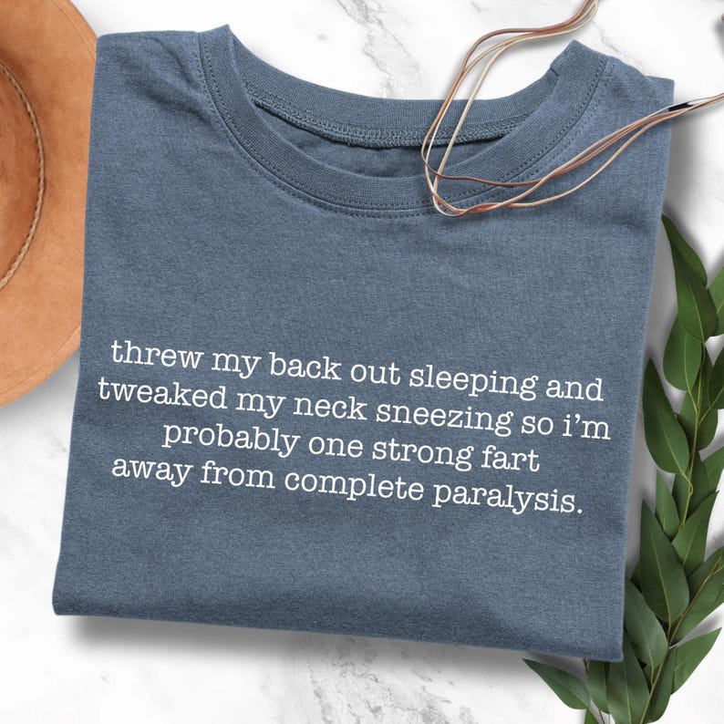May include: A blue t-shirt with white text that reads "threw my back out sleeping and tweaked my neck sneezing so i'm probably one strong fart away from complete paralysis."