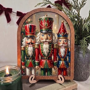 May include: An arched wooden frame displays a detailed illustration of three nutcracker figures. The central figure wears a red and green uniform, while the flanking figures wear green and blue uniforms respectively, all featuring white beards.