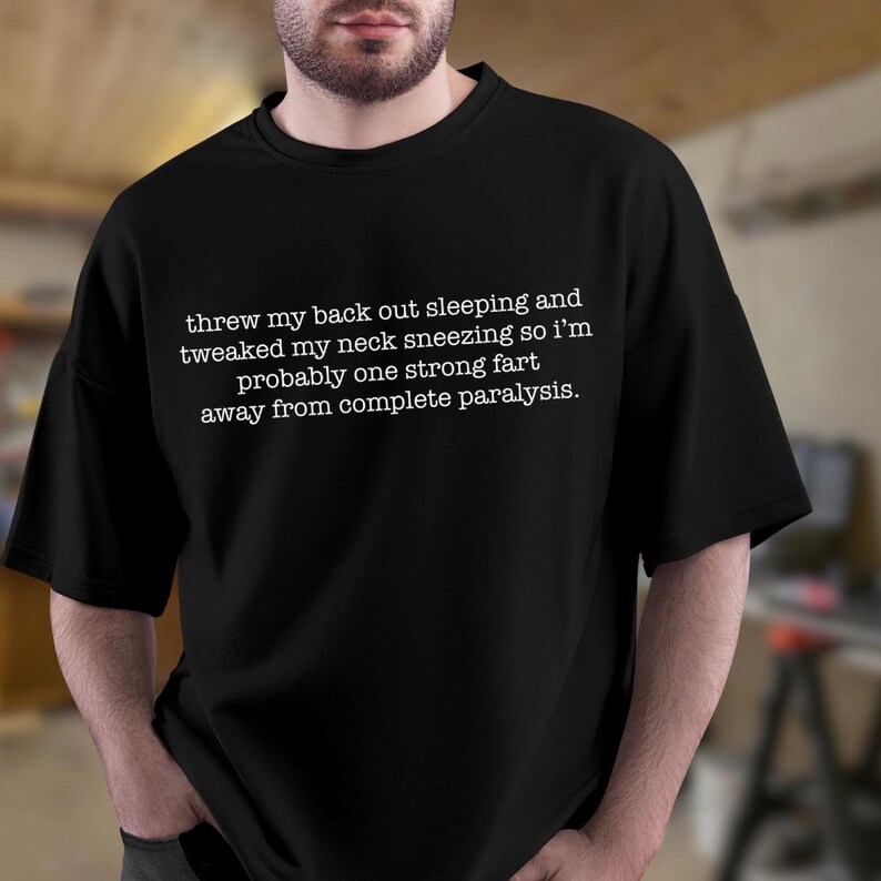May include: Black t-shirt with white text that reads "threw my back out sleeping and tweaked my neck sneezing so i'm probably one strong fart away from complete paralysis."