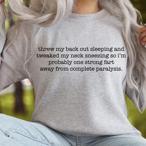 May include: Gray sweatshirt with a humorous saying: "threw my back out sleeping and tweaked my neck sneezing so i'm probably one strong fart away from complete paralysis."