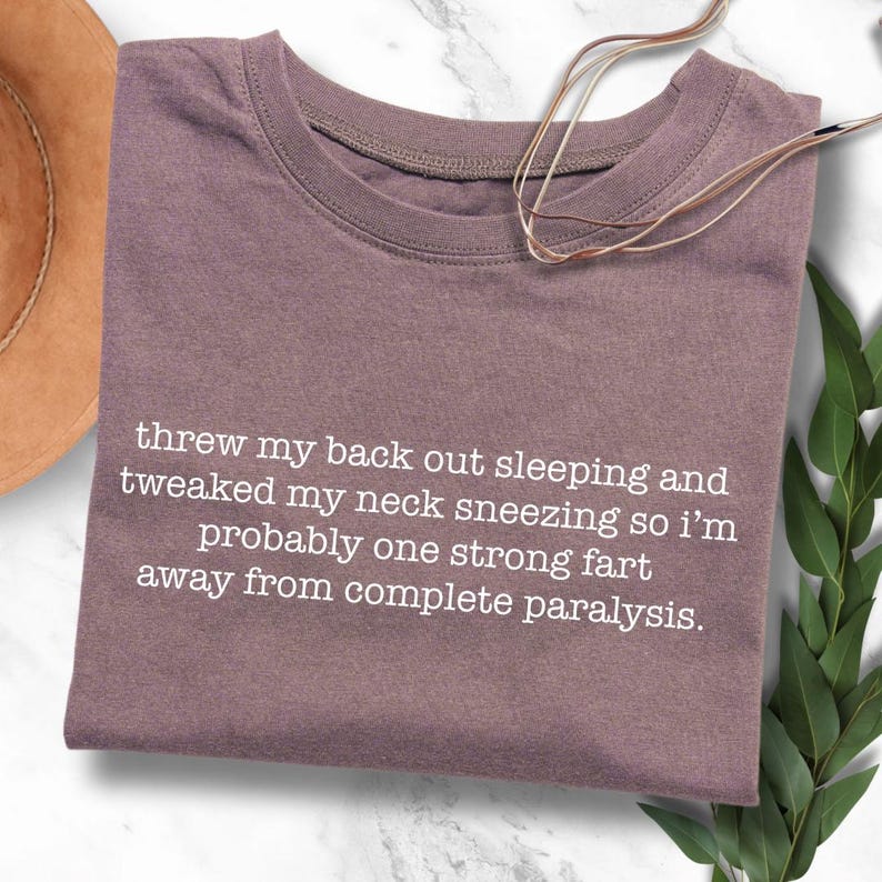 May include: A mauve colored t-shirt with white text that reads "threw my back out sleeping and tweaked my neck sneezing so I'm probably one strong fart away from complete paralysis."