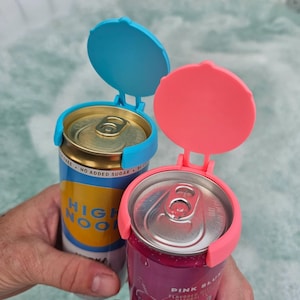 May include: Two aluminum beverage cans with protective can lids. One can is blue and white with the text "HIGH NOON". The other can is pink. The lids are blue and pink, and are attached to the cans with a hinge.