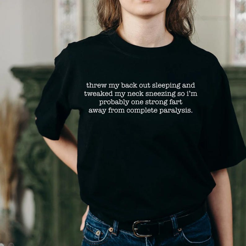 May include: Black t-shirt with white text that reads "threw my back out sleeping and tweaked my neck sneezing so I'm probably one strong fart away from complete paralysis."