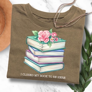 Puede incluir: Una camiseta marrón con una pila de libros y flores rosas. El texto "Cerré mi libro para estar aquí" está impreso en la camiseta.