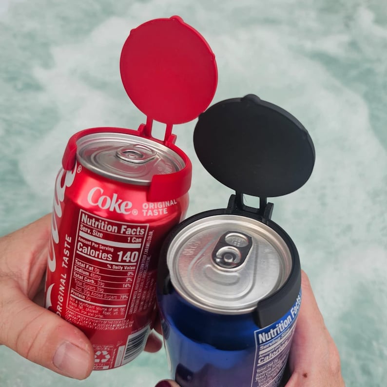 May include: Two soda cans, one red and one blue, each fitted with a hinged lid. The red can is labeled "Coke" with "Original Taste" text. The black and red lids are open, revealing the can openings. The image highlights a beverage accessory.