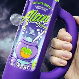 May include: A purple mug with a handle, designed to look like a can of "Witch's Brew" energy drink. The mug features a green rim, a cauldron graphic, and the word "Alan" in green. The background shows a moon and a night sky.