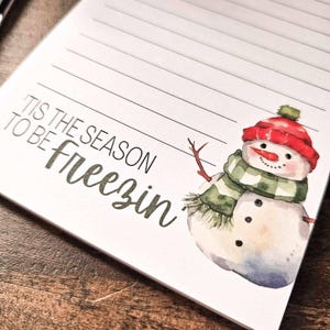 May include: A white notepad with a snowman illustration and the text "Tis the season to be freezin'" printed on the front.