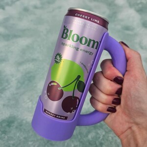 May include: A can of Bloom sparkling energy drink in a purple holder. The can is lavender with the words "Cherry Lime" and "Bloom" printed on it. The can features a graphic of cherries and a lime. The holder has a handle.