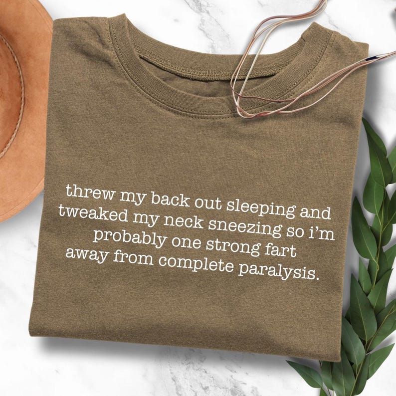 May include: A brown t-shirt with white text that reads "threw my back out sleeping and tweaked my neck sneezing so i'm probably one strong fart away from complete paralysis."