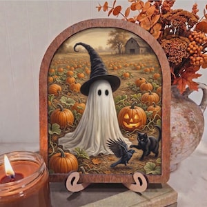 May include: A Halloween-themed wooden decoration featuring a ghost wearing a witch hat in a pumpkin patch. A black cat and crow are also depicted. The artwork is framed in a wooden arch and rests on a stand.