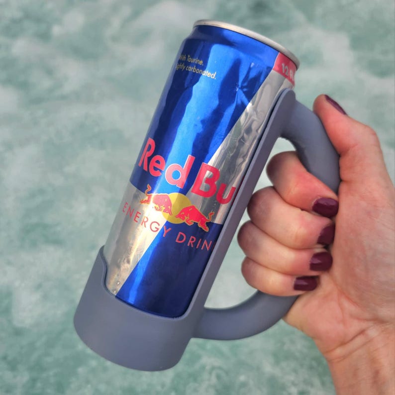 May include: A Red Bull energy drink can held in a gray silicone mug holder. The can has a blue and silver design with the Red Bull logo in red and yellow. The holder has a handle for easy gripping.