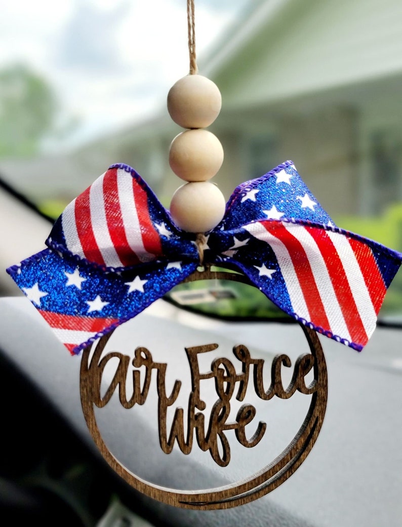 May include: A wooden car charm with a red, white, and blue ribbon and the words "Air Force Wife" in a circle. The charm is hanging from a string with three wooden beads.