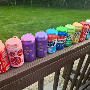 May include: A collection of beverage can-shaped containers with colorful lids. The cans feature various brand designs, including Coca-Cola, Pepsi, and Crown Royal Whisky. The containers are arranged on a wooden railing, showcasing a variety of colors and designs.