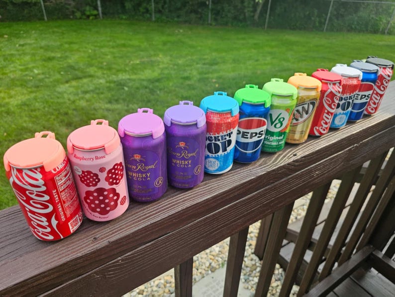 May include: A collection of beverage can-shaped containers with colorful lids. The cans feature various brand designs, including Coca-Cola, Pepsi, and Crown Royal Whisky. The containers are arranged on a wooden railing, showcasing a variety of colors and designs.
