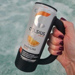 May include: A silver Celsius Sparkling Orange energy drink can held in a black handle. The can features orange and white text and a graphic of an orange slice. The text on the can includes "Celsius", "Live Fit", and "Sparkling Orange".