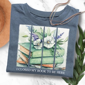 Könnte beinhalten: Ein blaues T-Shirt mit einem Aquarell-Design eines Stapels Bücher mit weißen Blumen und grünen Blättern. Der Text "Ich habe mein Buch geschlossen, um hier zu sein" ist unter dem Design gedruckt.