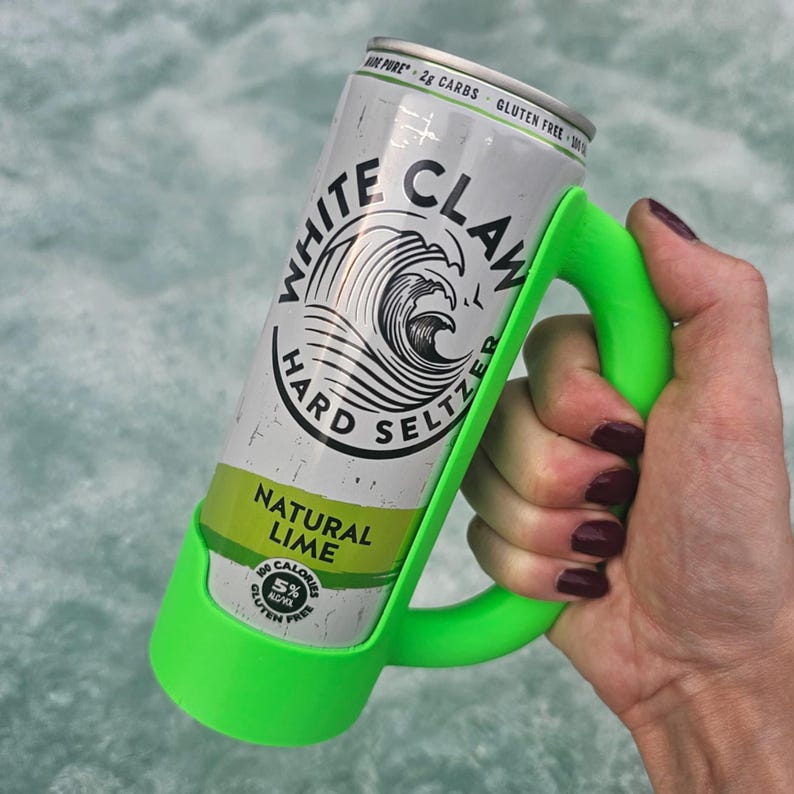 May include: A can of White Claw Hard Seltzer Natural Lime in a bright green holder. The can is silver with black and white text and a wave graphic. The holder has a handle for easy carrying.