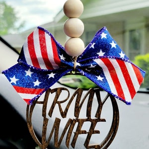 May include: A wooden car charm with the words "Army Wife" in a circle. The charm is decorated with a red, white, and blue bow with glitter stars. The bow is attached to a string of wooden beads.