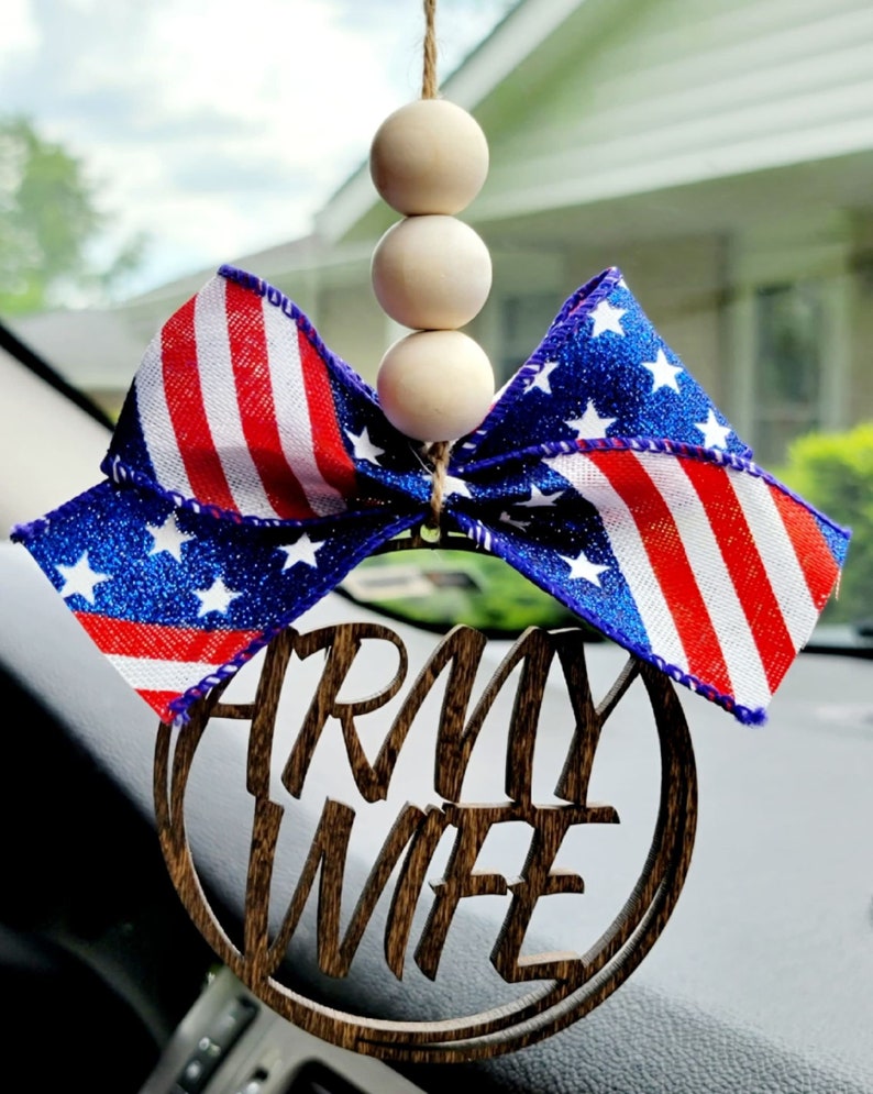 May include: A wooden car charm with the words "Army Wife" in a circle. The charm is decorated with a red, white, and blue bow with glitter stars. The bow is attached to a string of wooden beads.