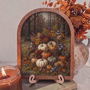 May include: An arched wooden frame displays a detailed painting of a harvest scene featuring various pumpkins, gourds, and autumn flowers nestled in a woven basket against a backdrop of a forest with muted fall foliage and earthy tones.