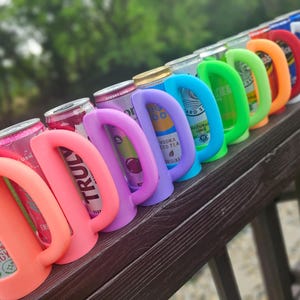 May include: A row of colorful can coolers in various shades, including pink, purple, blue, green, orange, red, white, and gray. Each cooler has a curved handle and is wrapped around a beverage can. The cans are visible through the cooler openings.