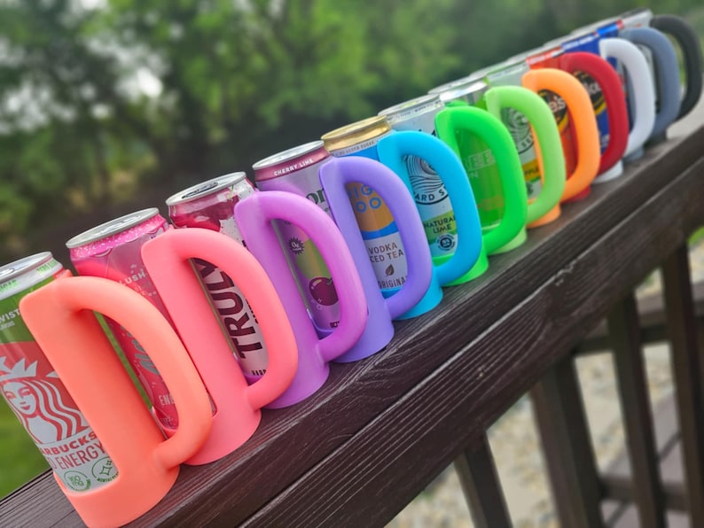 May include: A row of colorful can coolers in various shades, including pink, purple, blue, green, orange, red, white, and gray. Each cooler has a curved handle and is wrapped around a beverage can. The cans are visible through the cooler openings.